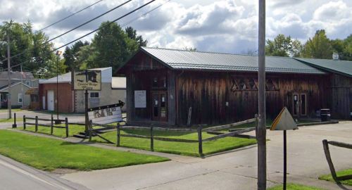 A rustic wooden building with a green roof, featuring a sign, surrounded by grass and a fence on a cloudy day.