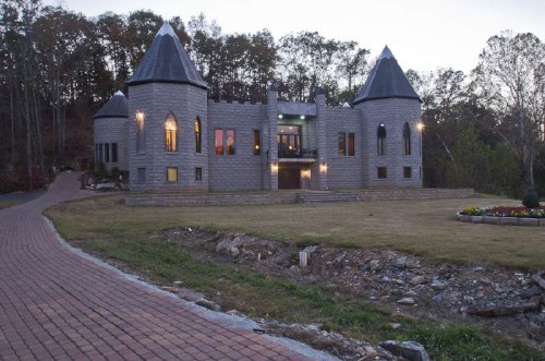 A stone castle with turreted towers, set in a wooded area, with a paved driveway leading up to it at dusk.