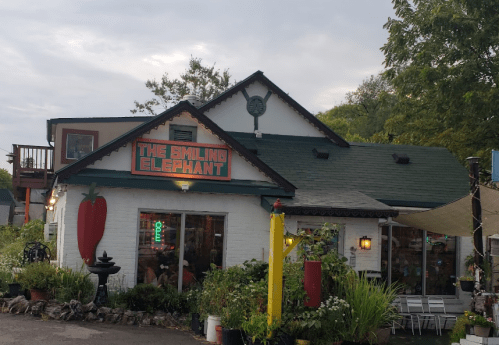 A charming restaurant called "The Smiling Elephant" with a colorful sign, surrounded by greenery and outdoor seating.