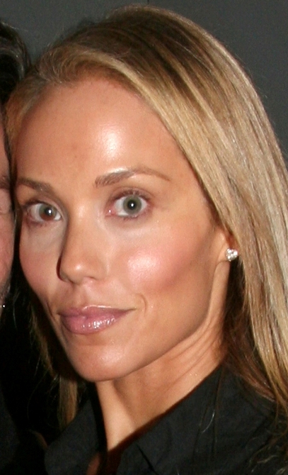 A close-up portrait of a woman with long blonde hair, wearing a black top and subtle makeup, smiling softly.