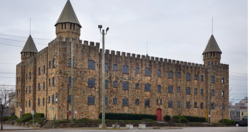 A large stone castle-like building with towers, surrounded by power lines and greenery, under a cloudy sky.