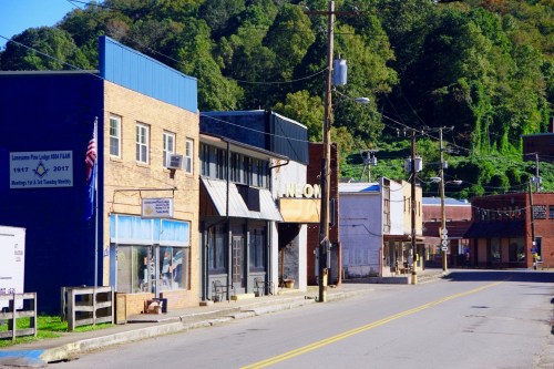 A quiet street in a small town, featuring brick buildings, a blue storefront, and lush green hills in the background.