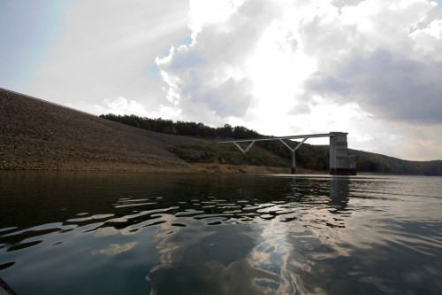 A serene view of a dam with a tower, reflecting clouds and trees in the calm water below.