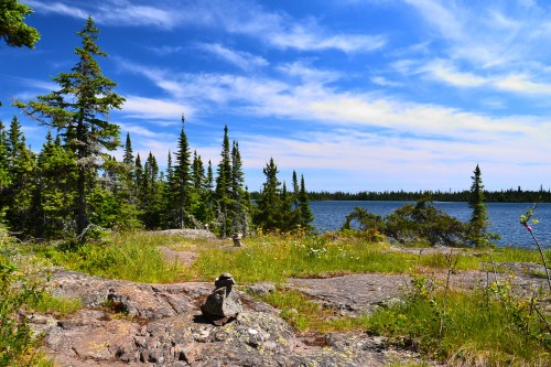 A serene lakeside scene with lush green trees, rocky terrain, and a clear blue sky dotted with clouds.