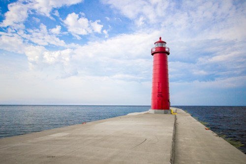A tall red lighthouse stands on a concrete pier, overlooking calm waters under a partly cloudy sky.