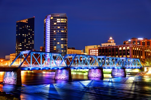 A brightly lit bridge spans a river at night, with modern buildings and a city skyline in the background.