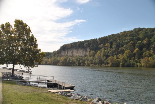 A serene river scene with a rocky shore, a dock, and a forested cliff under a partly cloudy sky.