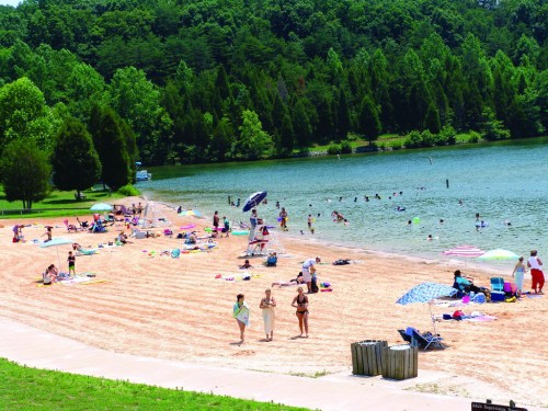 A sandy beach with people sunbathing, swimming, and relaxing by a lake, surrounded by green trees and hills.