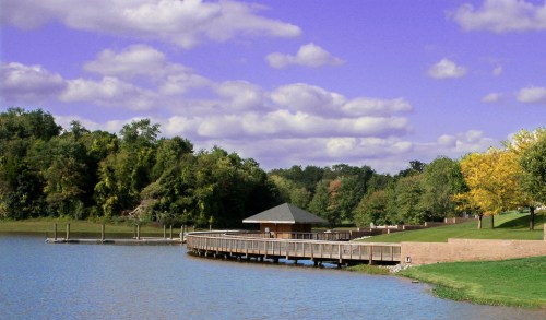 A serene lake scene with a wooden dock, a small pavilion, and colorful trees under a blue sky with fluffy clouds.