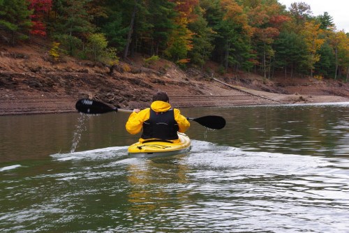 A person in a yellow kayak paddles on a calm lake surrounded by autumn-colored trees.