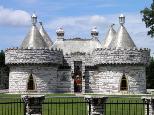 A whimsical stone castle with conical towers and decorative elements, surrounded by green grass and a black fence.