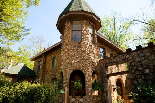 A unique stone house with a turret, surrounded by greenery and trees under a clear blue sky.