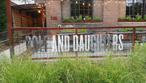 Sign reading "Ron and Daughters" in front of a brick building, surrounded by greenery and outdoor seating.