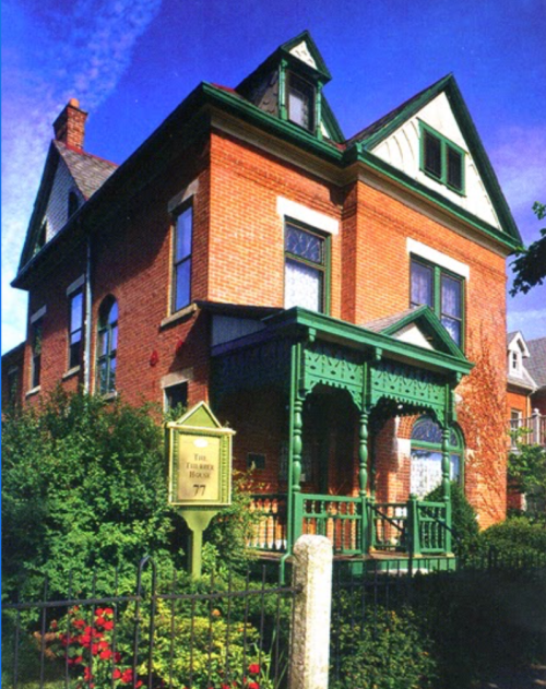 A historic brick house with green trim and a porch, surrounded by greenery and a white fence.