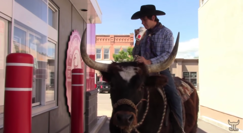 A person in a cowboy hat rides a large bull at a drive-thru, holding a drink and smiling.