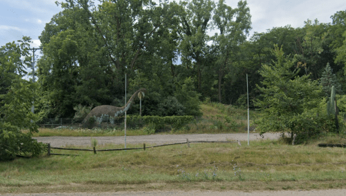 A large dinosaur statue surrounded by trees and greenery in a rural setting.