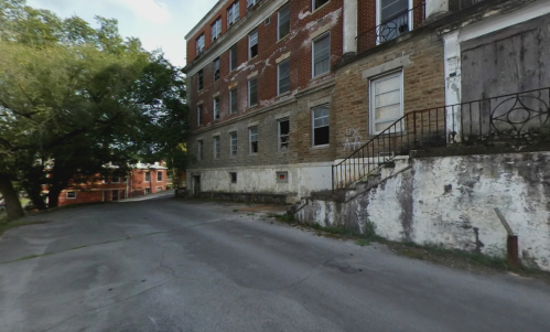 Abandoned building with brick and stone walls, broken windows, and a cracked staircase, set on a quiet street.