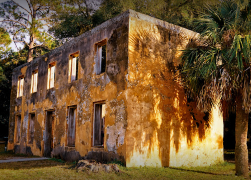 Abandoned yellow building with weathered walls and windows, surrounded by trees and grass, casting long shadows.