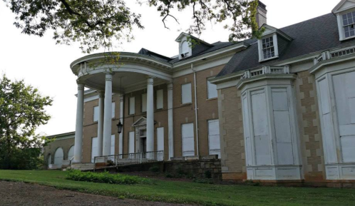 Historic mansion with white columns and boarded windows, set in a grassy area under a clear sky.
