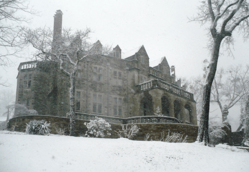 A large, historic stone mansion covered in snow during a winter storm, surrounded by bare trees.