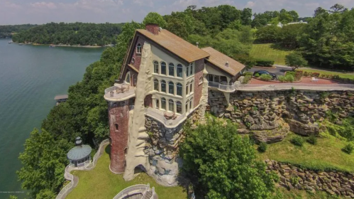 A unique house built into a rocky cliff, overlooking a lake, surrounded by lush greenery and a clear blue sky.