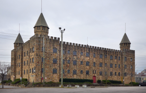 A large stone castle-like building with towers, featuring multiple windows and a red door, set against a cloudy sky.