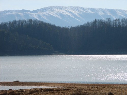 A serene lake reflects sunlight, with snow-capped mountains and trees in the background under a clear blue sky.