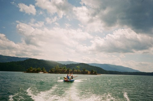 A group of people on a boat enjoying a sunny day on a lake, with mountains and clouds in the background.