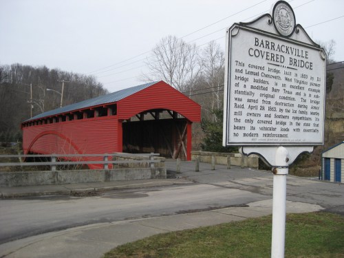 A red covered bridge with a sign nearby, set against a hilly landscape and a clear sky.