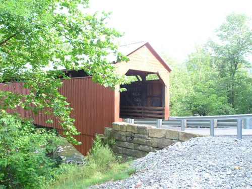 A red covered bridge surrounded by greenery, with a gravel path and guardrail nearby.