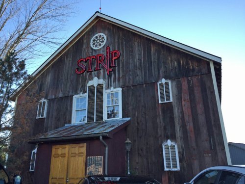 A rustic barn with a neon "STRIP" sign, featuring wooden siding and white shutters under a clear blue sky.