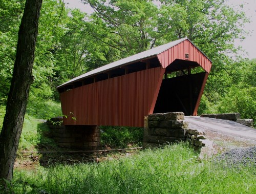 A red covered bridge spans a small creek, surrounded by lush green trees and grass.