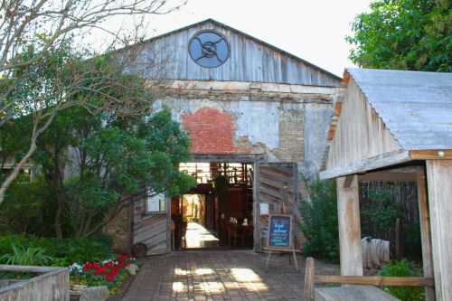 A rustic barn entrance with weathered wood, greenery, and a sign, inviting visitors into a cozy interior.