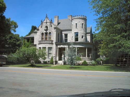 A large, ornate stone mansion with a turret, surrounded by greenery and a clear blue sky.