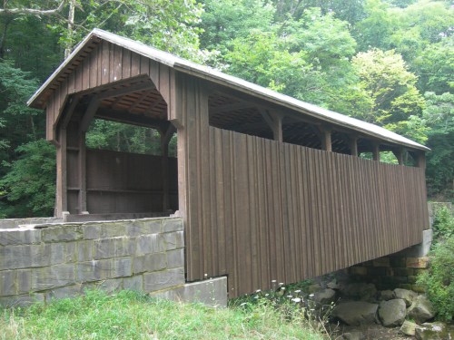 A wooden covered bridge spans a small stream, surrounded by lush greenery and trees.