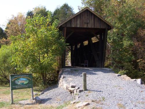 A wooden covered bridge surrounded by trees, with a gravel path and a sign nearby.