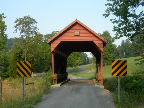 A red covered bridge spans a rural road, flanked by warning signs, surrounded by green trees and fields.