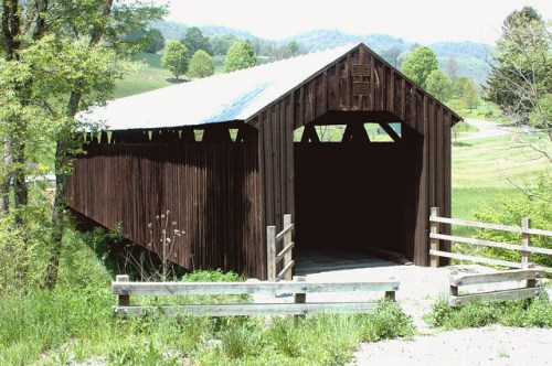 A rustic wooden covered bridge surrounded by greenery and rolling hills on a sunny day.