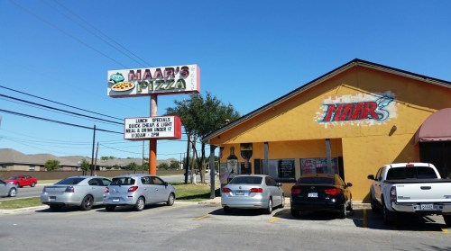 A colorful pizza restaurant with a sign advertising lunch specials, surrounded by parked cars under a clear blue sky.