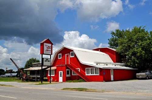 A red barn-style building with a sign, surrounded by greenery and a cloudy sky, located by a road.