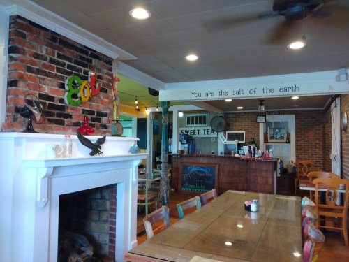 Cozy restaurant interior with a brick wall, colorful decor, and a large wooden table. "You are the salt of the earth" sign visible.