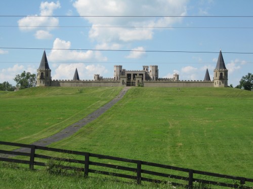 A large castle with towers sits atop a green hill, surrounded by a grassy landscape and a black fence in the foreground.