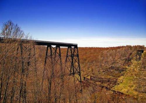 A tall, abandoned railway trestle bridge spans a wooded area with autumn foliage under a clear blue sky.
