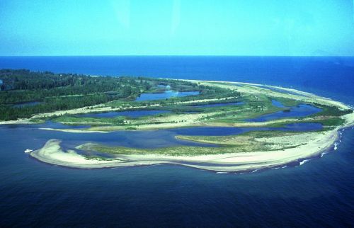 Aerial view of a coastal island with sandy shores, lush greenery, and winding waterways under a clear blue sky.
