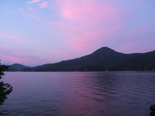 A serene lake at sunset, with soft pink skies and mountains reflecting on the water's surface.