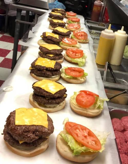 A row of burger patties with cheese, lettuce, and tomato on buns, ready for assembly in a kitchen.