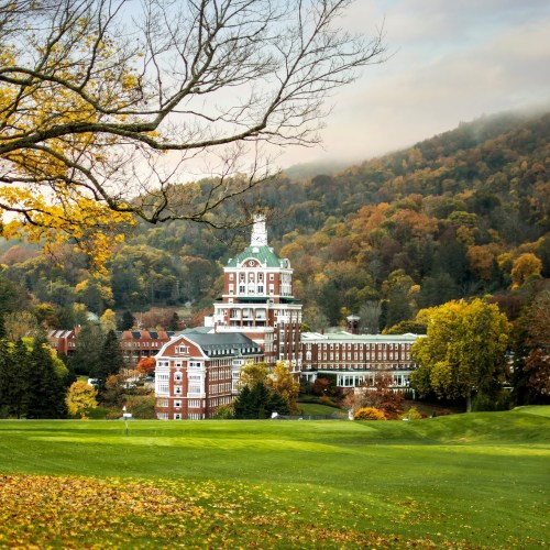A grand hotel surrounded by autumn foliage and rolling hills, with a cloudy sky above.