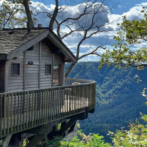 A wooden cabin with a wraparound deck overlooks a lush green valley and blue sky with scattered clouds.