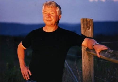 A man in a black shirt leans against a wooden fence, smiling softly in a scenic outdoor setting at dusk.