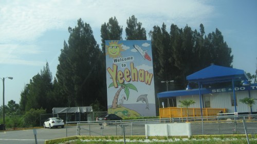A colorful sign reading "Welcome to Yeehaw" with a sun, dolphin, and tropical theme, surrounded by trees.